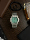 Silver men's luxury watch with green dial on wood surface, surrounded by wallet, bracelet, and cologne