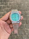 Luxury men's silver wristwatch with turquoise dial, held in hand on stone pavement background.